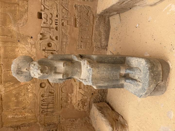 Granite statue of the lioness goddess Sekhmet seated against a wall of hieroglyphs in a sandy courtyard.