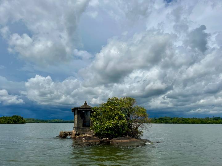 Small shrine and trees on a rock outcrop in a wide lake under dramatic clouds.