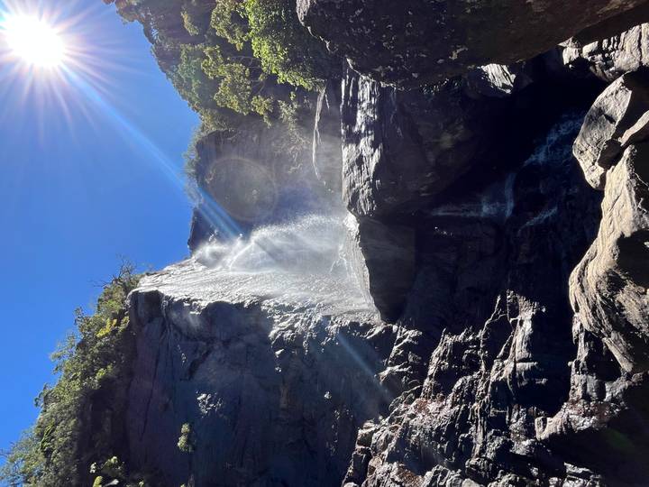 Tall waterfall cascades down rugged rocks with sun flare against a clear sky.