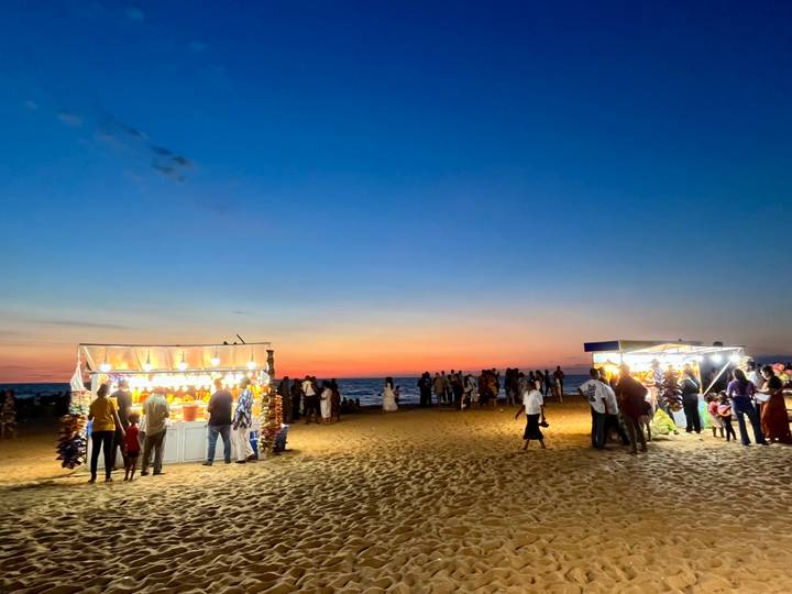 Evening beach scene with brightly lit snack stalls and crowds against a colorful sky.