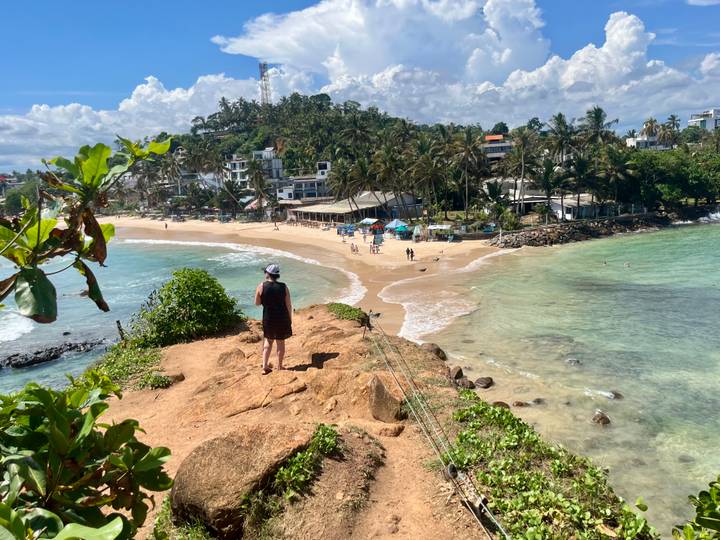 Person stands on a rocky bluff overlooking a curved sandy beach and turquoise sea.