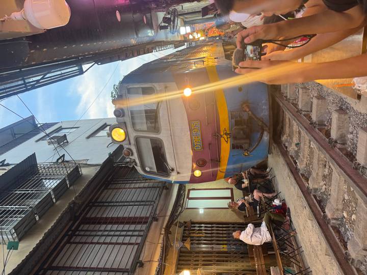 Close-up of locomotive squeezing past outdoor café tables as onlookers snap photos on Hanoi Train Street.