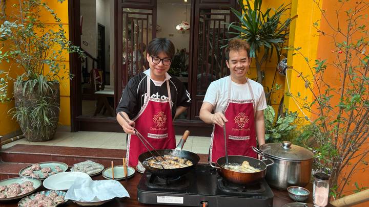 Two young men in aprons frying food on outdoor stoves during a Vietnamese cooking class.