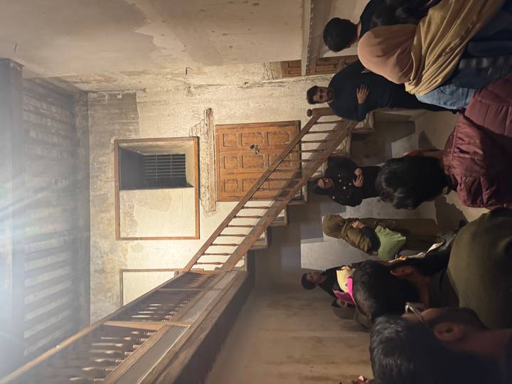 Visitors listen to a guide inside an aged stone building with wooden stairs and faded walls.