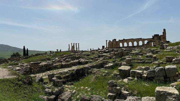 Wide view of ancient stone ruins scattered across a green grassy hillside under a bright blue sky.