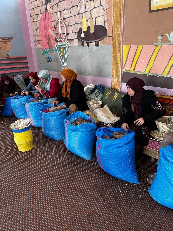 Five women sit in a row indoors sorting nuts into bright blue sacks as part of a traditional cooperative.