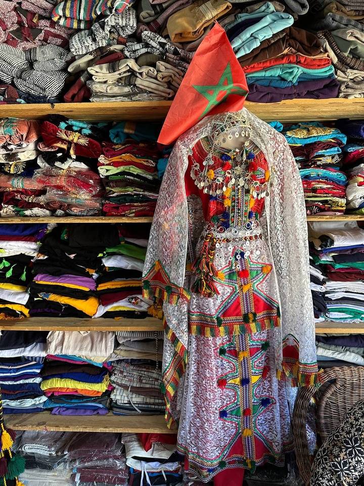 Colorful traditional Berber dress adorned with jewelry displayed in front of neatly stacked fabrics.