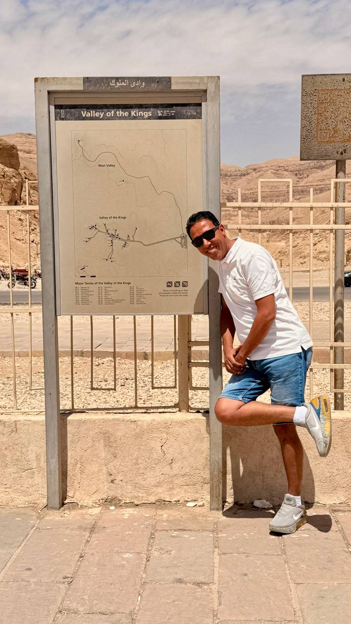 Traveler in white polo smiles beside an information map for the Valley of the Kings in a desert setting.