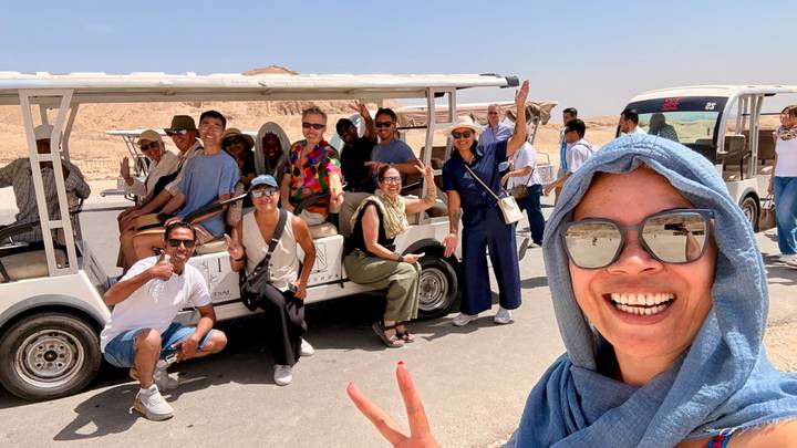 Large tour group waves from open electric shuttles in a sun-baked desert archaeological area.