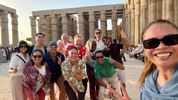 Cheerful tour group poses with victory signs among towering sandstone columns of Luxor Temple at sunset light.