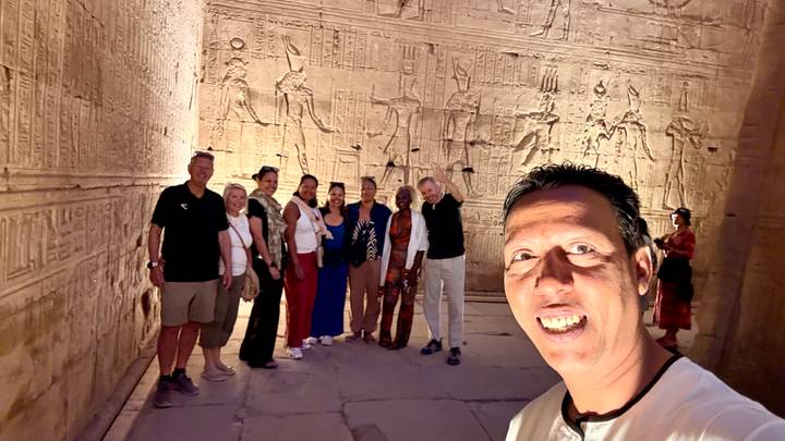 Small group stands inside an ornately carved ancient temple corridor illuminated by warm artificial light.
