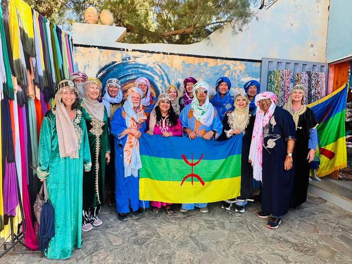 Tour group dressed in colourful Berber attire proudly holds an Amazigh flag inside a textile shop.
