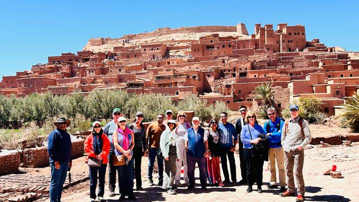 Large tour group stands before the fortified clay village of Ait Benhaddou under clear skies.