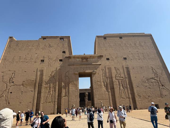 Massive sandstone entrance walls of Edfu Temple adorned with ancient carvings, crowds gathered below.