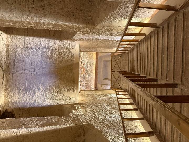 Dimly lit wooden staircase descending into the interior of an ancient Egyptian tomb with carved walls.