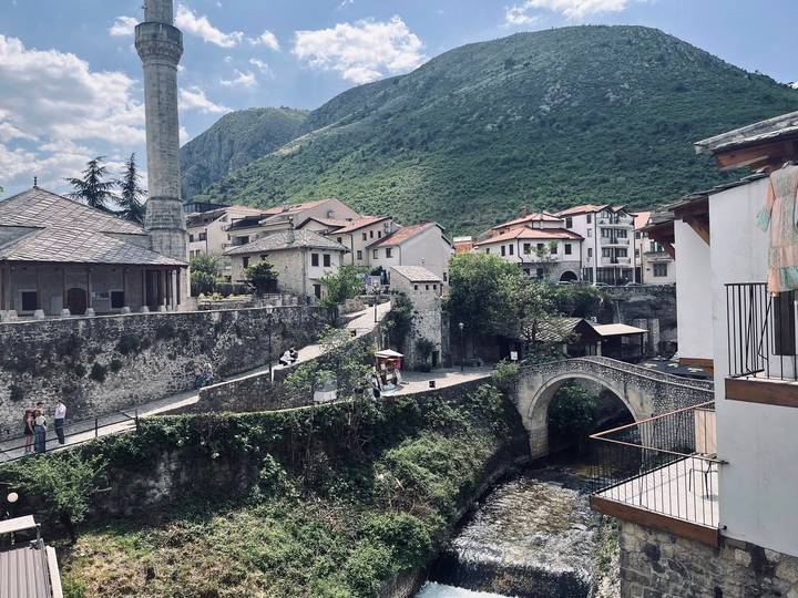 Stone arch bridge spans a narrow river below hillside homes and a minaret, backed by green mountains.