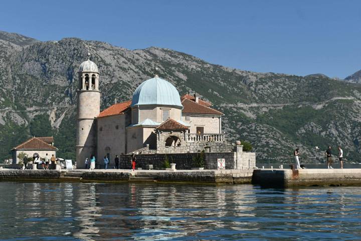 Stone church of Our Lady of the Rocks sits on an islet amid calm bay waters backed by rugged mountains.