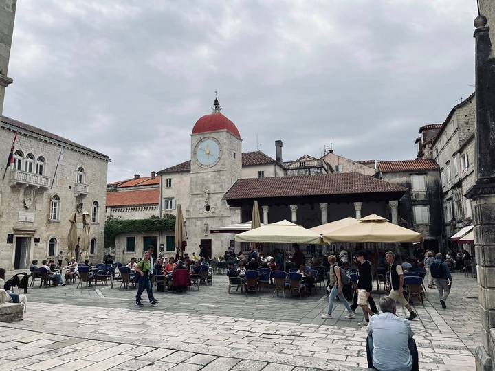 Lively town square in Trogir with a red-topped clock tower and café umbrellas.