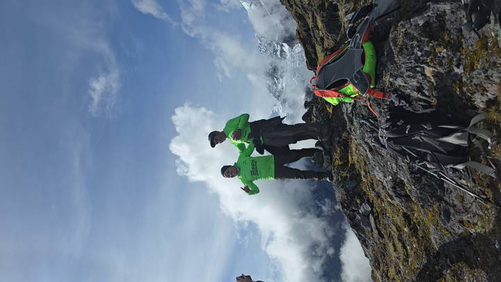 Two trekkers in bright shirts celebrate atop a rocky Andean ridge with snowy peaks behind.
