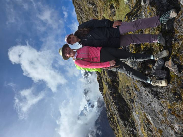 Two hikers smile on a rocky slope with glaciated Andean mountains rising into the clouds.