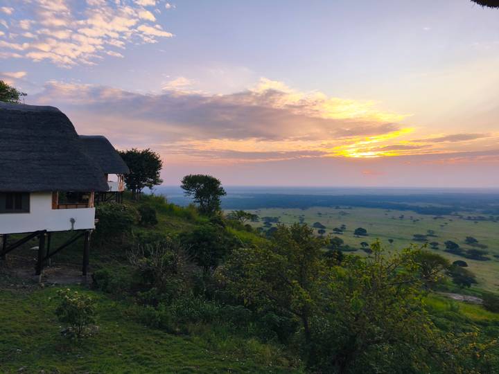Elevated lodge with thatched roof perched on a verdant hillside overlooking a vast plain at sunrise.