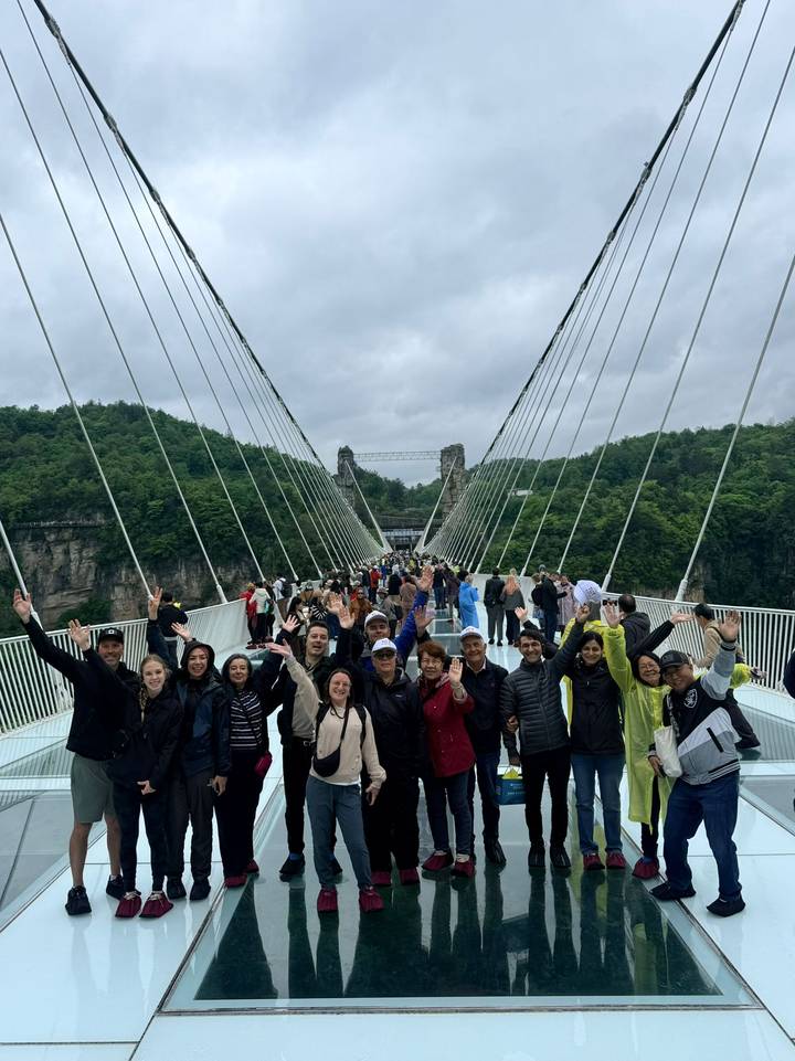 Tourists cheer on a long glass suspension bridge spanning a deep gorge with lush cliffs.