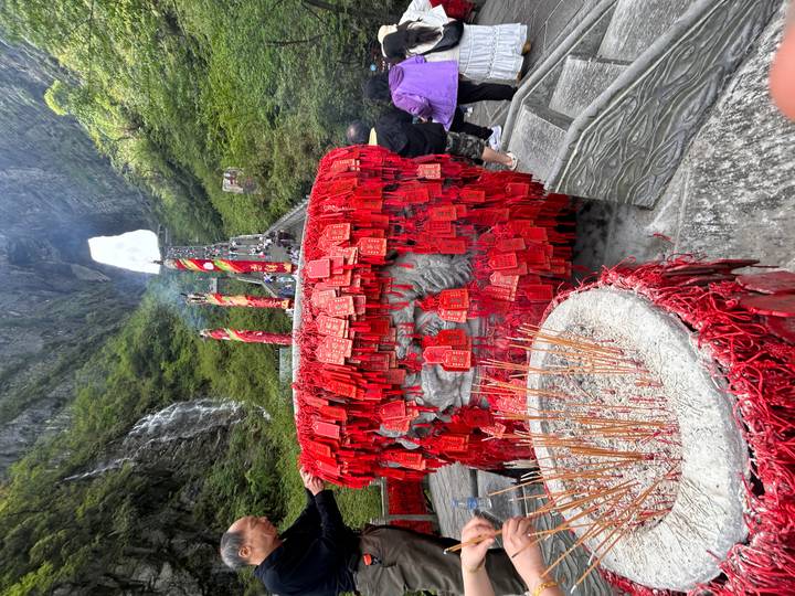 Incense burner and red wish plaques with smoke rising at a mountain shrine, towering arch visible in background.