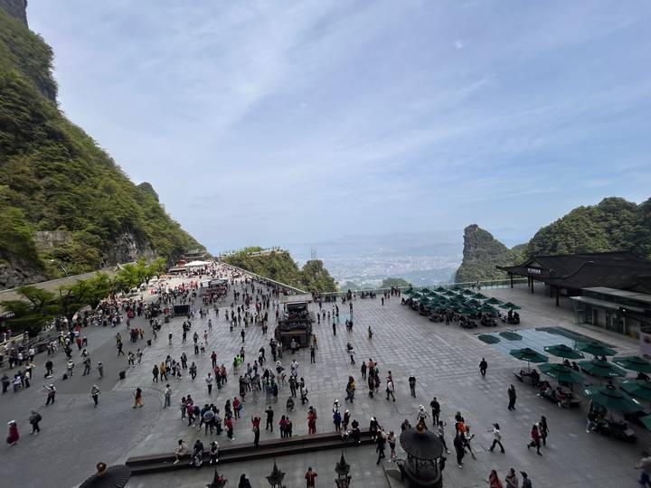Wide plaza atop mountain ridge filled with visitors, offering panoramic views of distant peaks and city below.