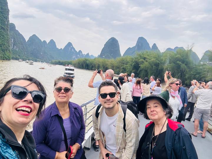 Travelers on a river cruise deck with karst mountains and bamboo groves lining the waterway.