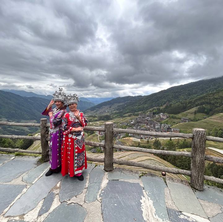 Two women in ornate ethnic dress pose on a terrace overlooking tiered rice terraces and mountain village.