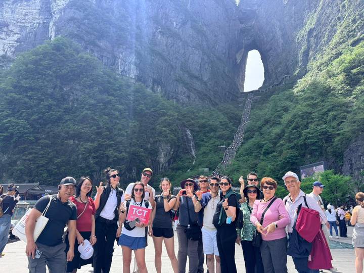 Tour group posing below Tianmen Mountain’s massive rock arch with steep staircase behind.