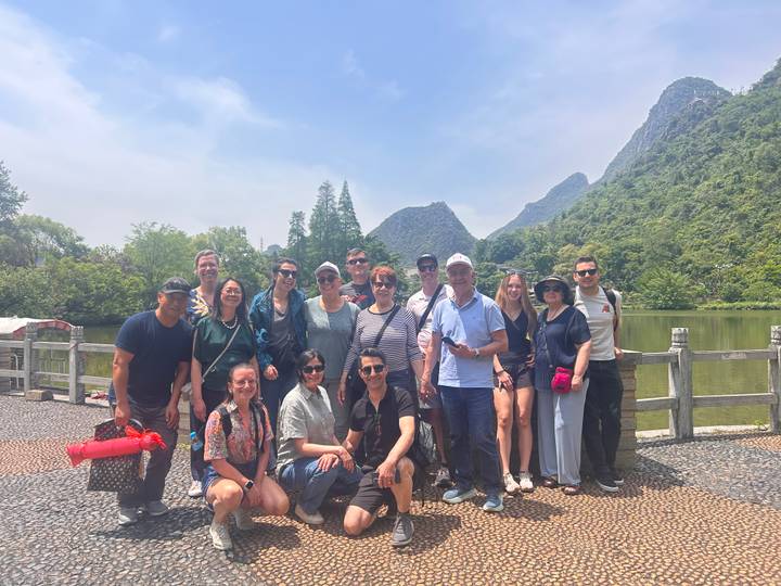 Smiling tour group poses beside a tranquil lake with jagged karst peaks and greenery under a blue sky.
