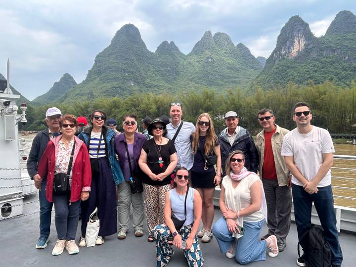 Tourists stand on a riverboat deck with lush karst mountains rising behind them.