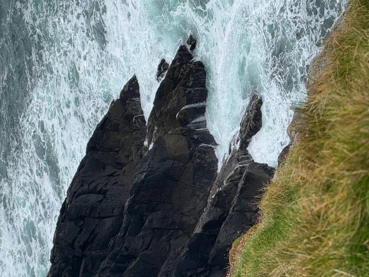 Wild Atlantic waves crash against dark jagged rocks below a grassy cliff edge.
