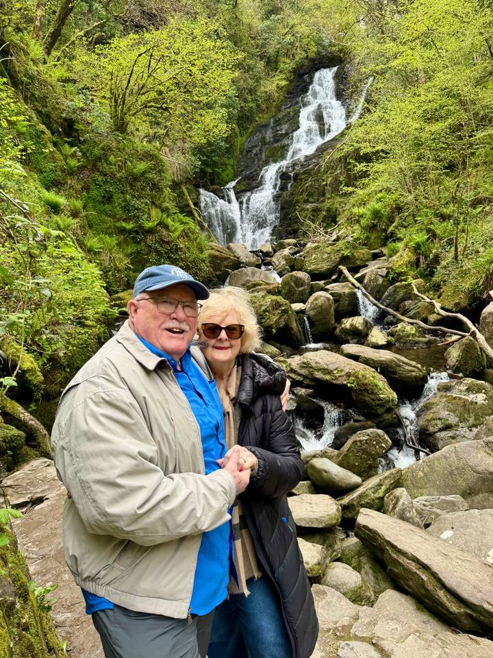 Smiling couple poses in front of mossy rocks and a small forest waterfall.