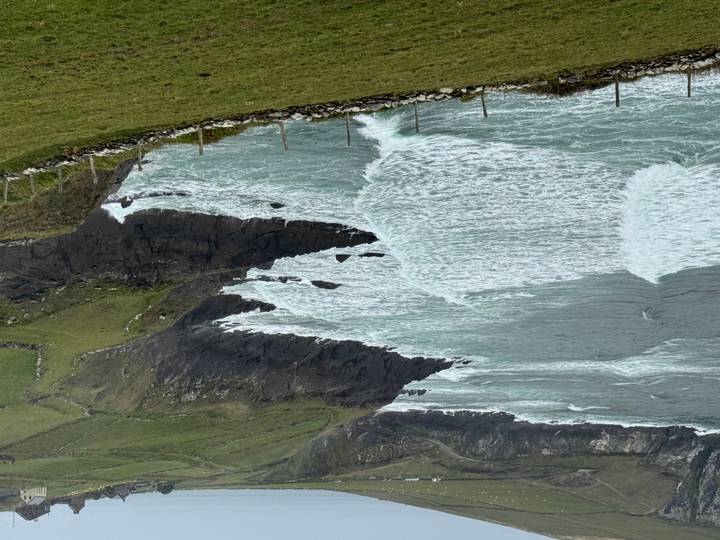 Wide view of rolling Atlantic waves hitting rugged Irish cliffs and green pastureland.