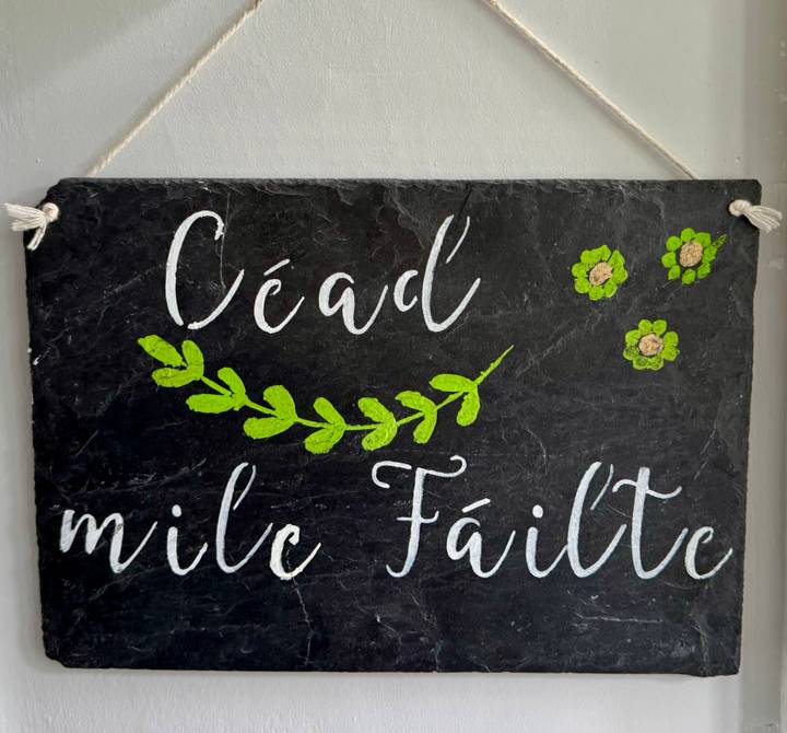 Slate welcome sign painted in Gaelic with green leaf design hanging on a wall.