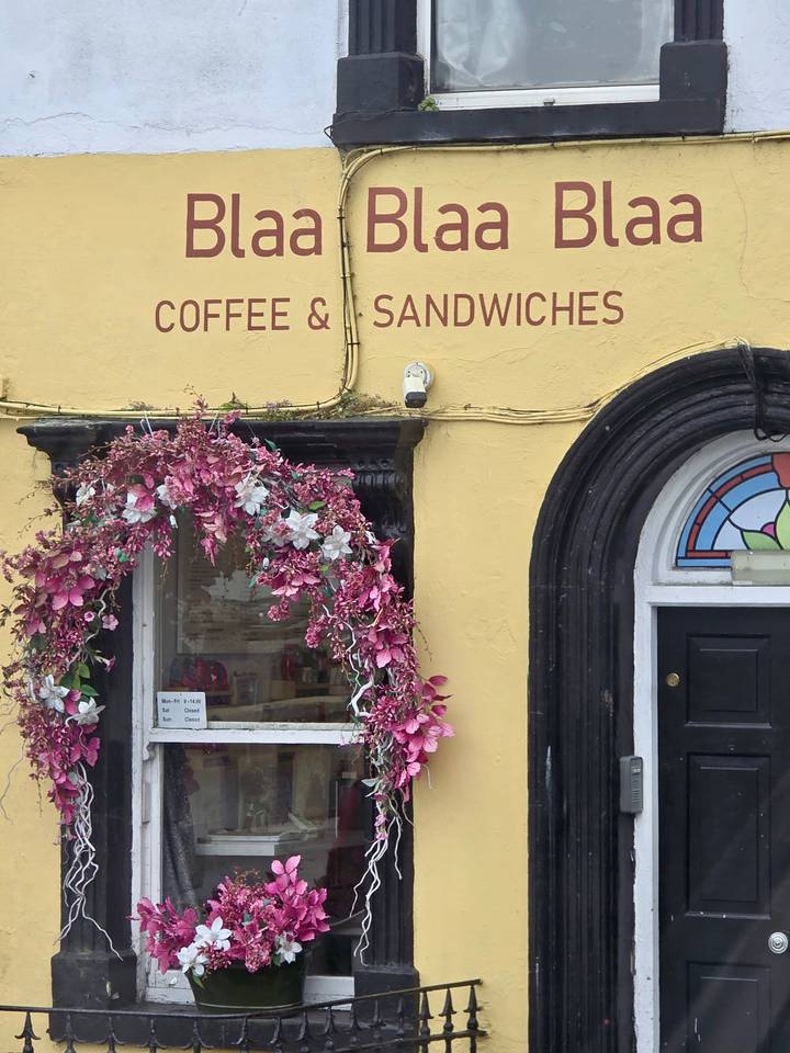 Yellow storefront with arched black trim, pink floral decoration and sign for coffee and sandwiches.