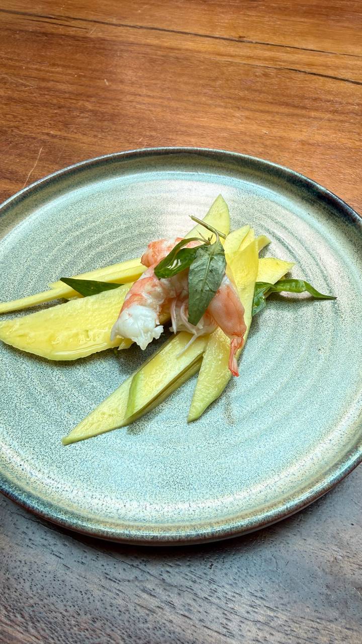 Plate with sliced mango topped with a single prawn and basil leaves on a textured dish.