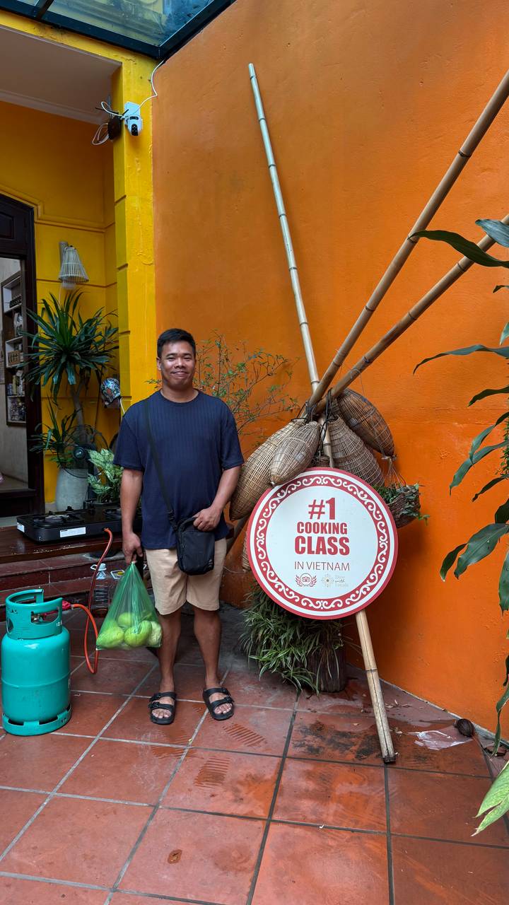 Smiling participant stands beside a bright orange wall and a sign advertising the cooking class.
