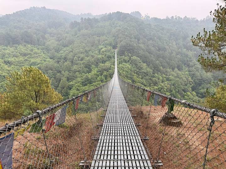 Long metal suspension bridge with prayer flags stretching across a deep green forested valley.