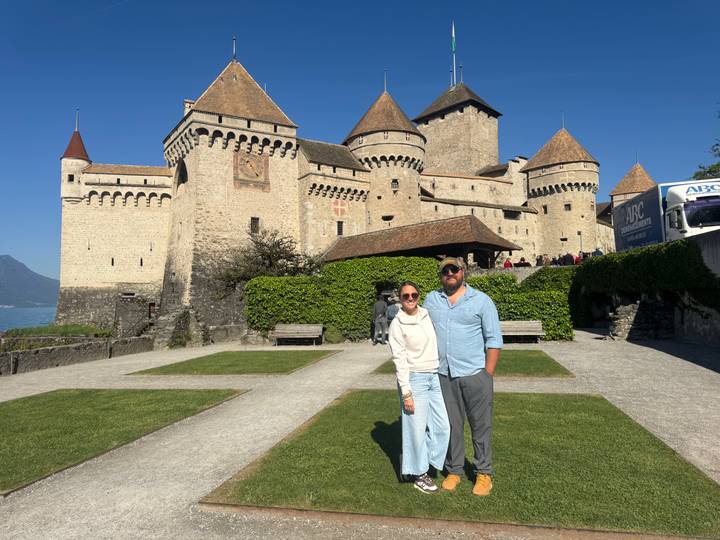 Couple posing in front of medieval lakeside Château de Chillon with clear blue sky.