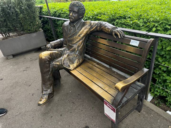 Bronze statue of a seated man on a bench set beside manicured hedges.