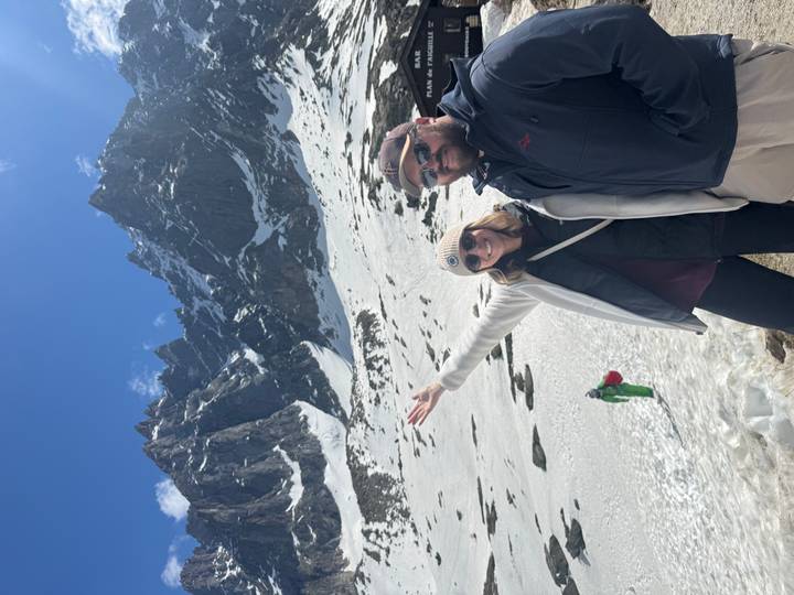 Smiling couple in snowy alpine terrain with jagged peaks rising behind them.