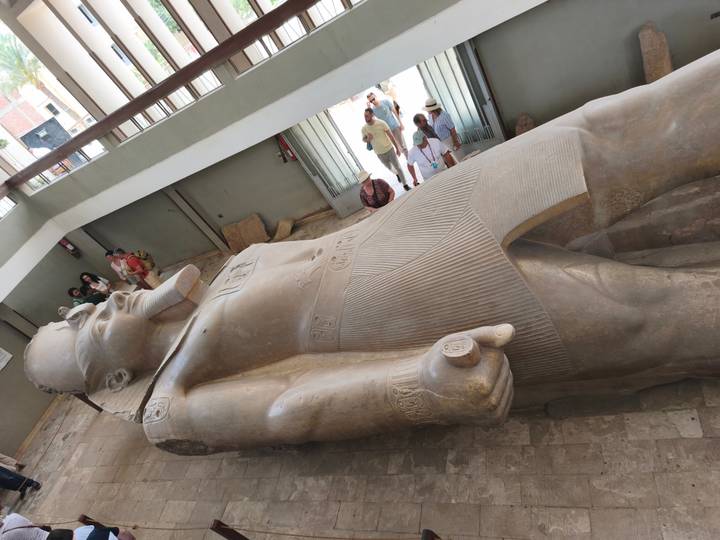 Massive reclining statue of Ramses II in indoor museum with visitors looking on.
