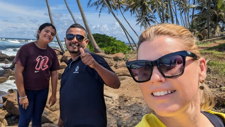 Three travellers pose by a rocky tropical coastline dotted with palm trees beneath a bright blue sky.
