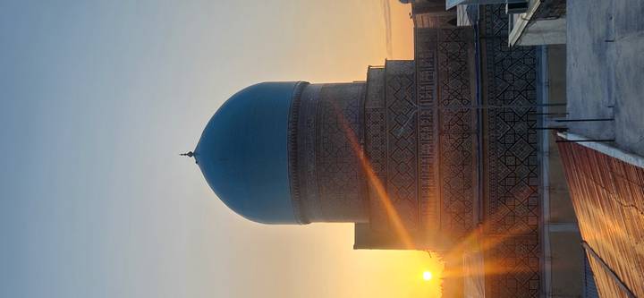 Blue-tiled domed mausoleum glowing in the warm light of sunset with sunburst on horizon.