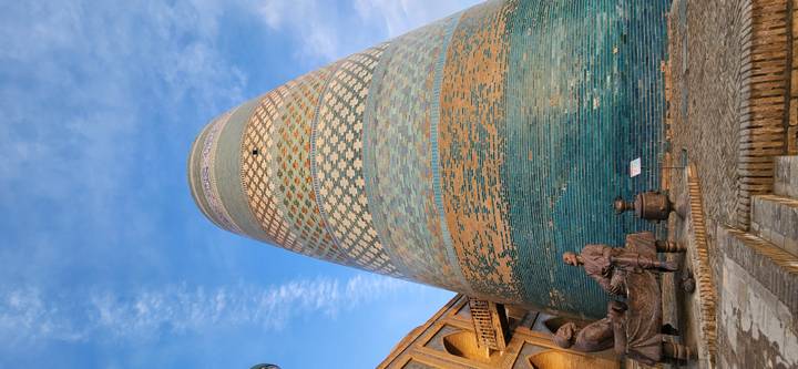 Massive blue-green tiled Kalta Minor minaret towers above brick buildings with statues at its base.