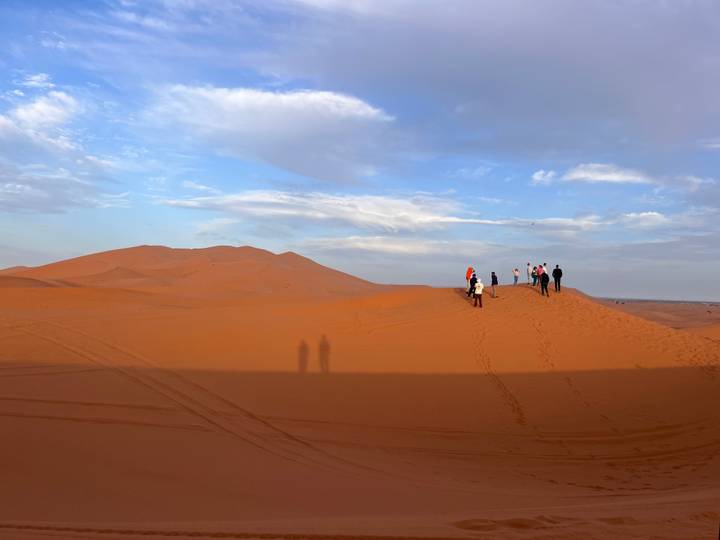 Travellers walk along the crest of vast orange sand dunes at golden hour casting long shadows across the Sahara.