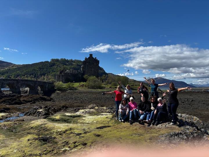 Small tour group poses happily in front of Eilean Donan Castle with mountains behind.